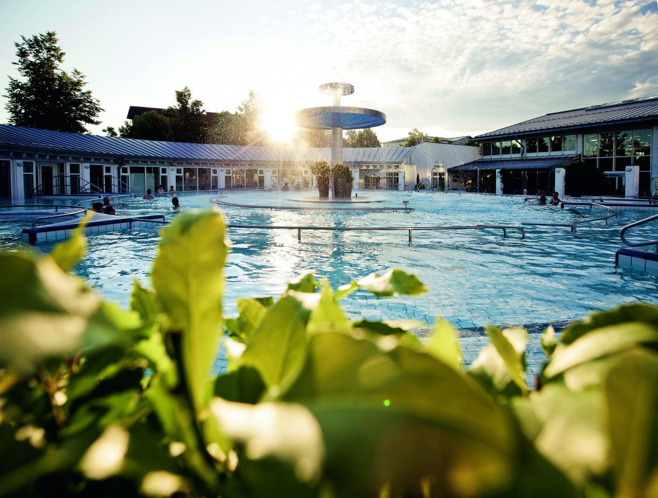 Ein Außenbereich eines Schwimmbades mit einem großen Wasserfall in der Mitte. Das Wasser glitzert unter der Sonne, während um den Pool herum Pflanzen und Bäume wachsen. Menschen entspannen sich im Wasser.