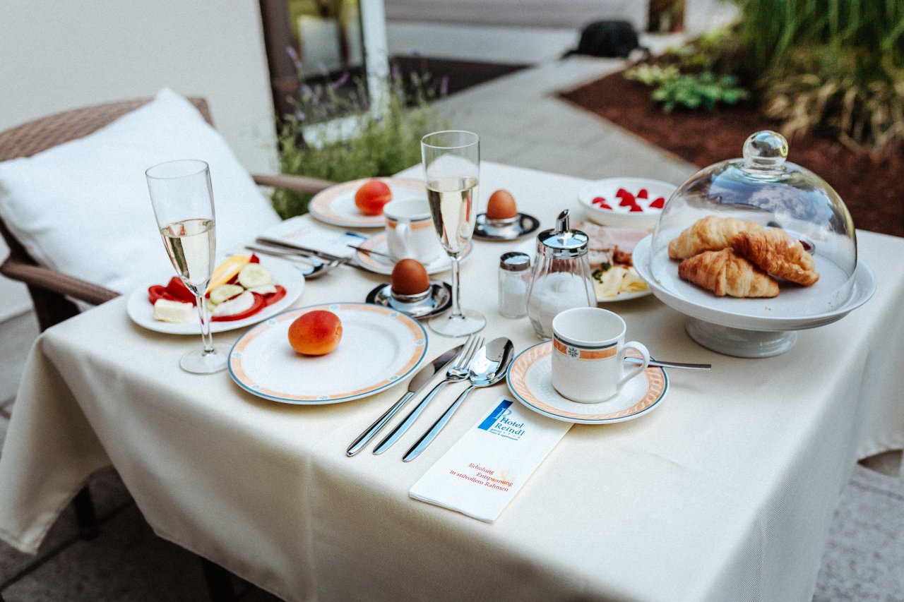Ein schön gedeckter Tisch im Freien mit verschiedenen Frühstücksgerichten. Darauf sind frisches Obst, Eier im Eierbecher, Croissants, Sektgläser und eine Kaffeetasse zu sehen. Die Tischdecke ist beige und das Ambiente wirkt einladend.