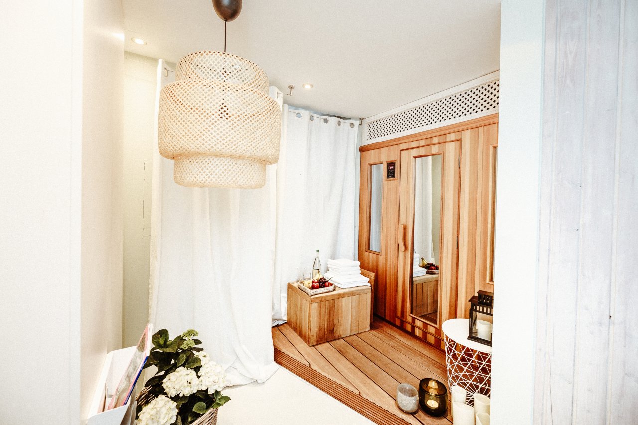 Ein eleganter Spa-Bereich mit einem Holzboden, einer Sauna aus Holz, einem großen Wäschekorb und einem dekorativen Raumteiler. Am Eingang hängen eine stilvolle Korbpendelleuchte und frische Blumen.