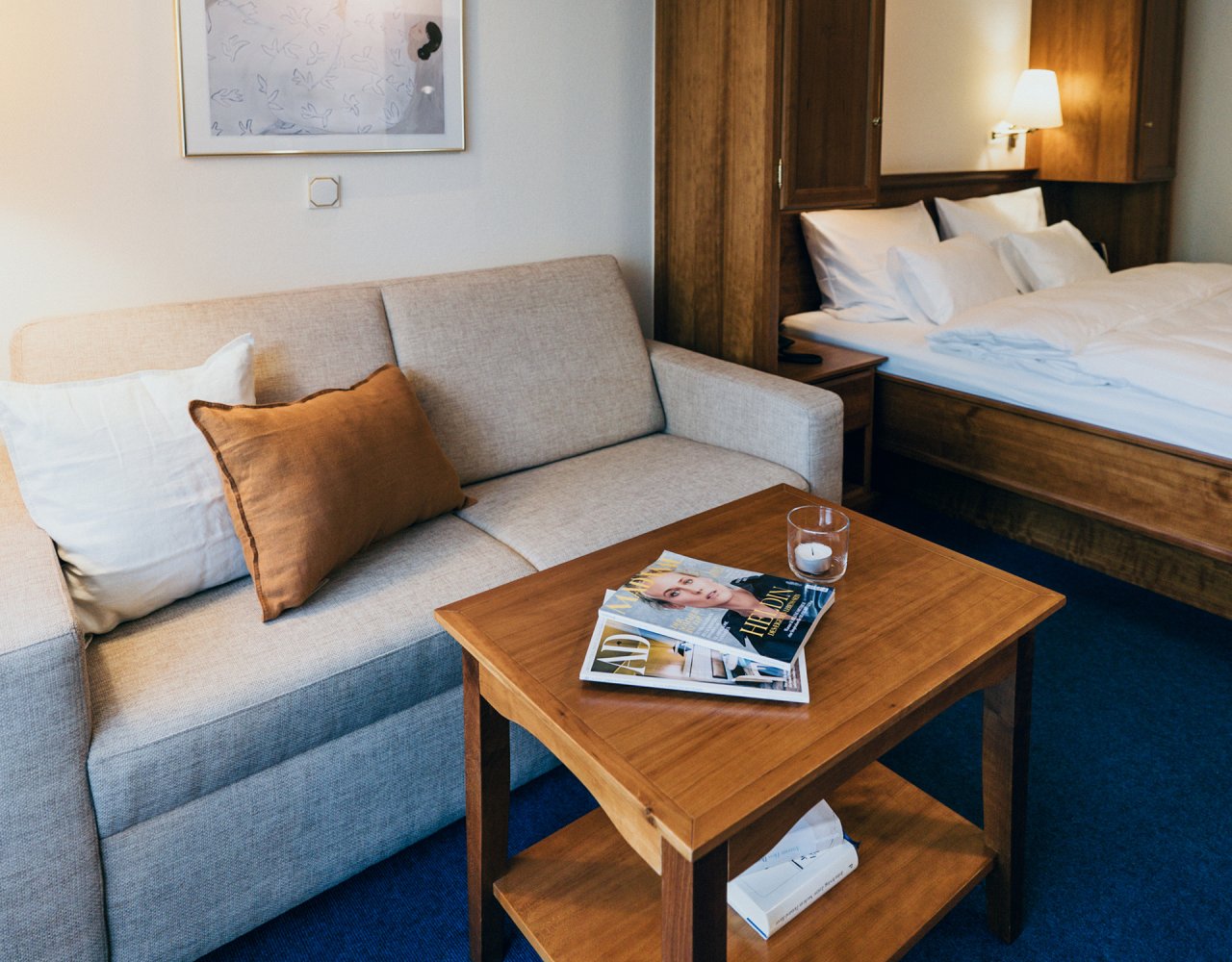 Ein heller Hotelzimmer mit einem grauen Sofa, einem Holztisch, einem Magazin auf dem Tisch und einem Bett im Hintergrund. Es gibt eine ruhige Atmosphäre mit einem warmen Licht.